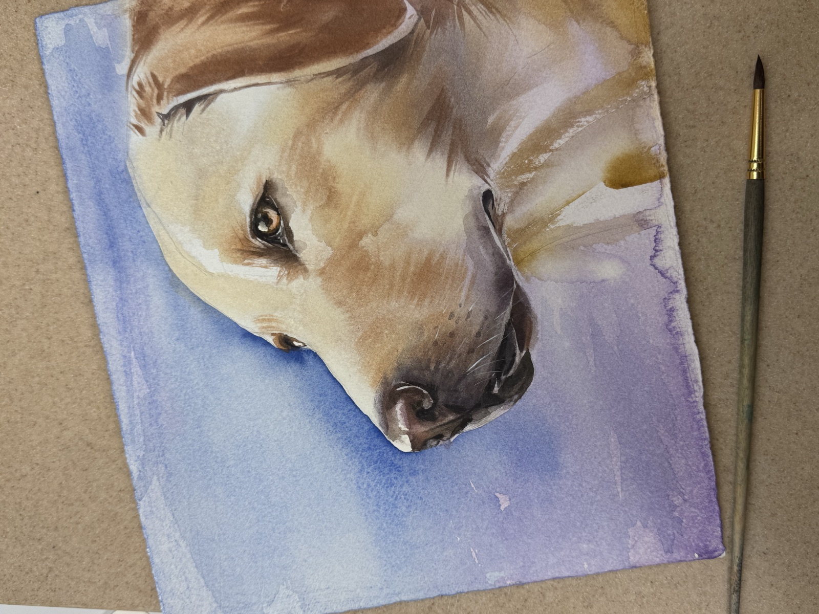 Watercolor portrait of a golden Labrador on paper beside a paintbrush